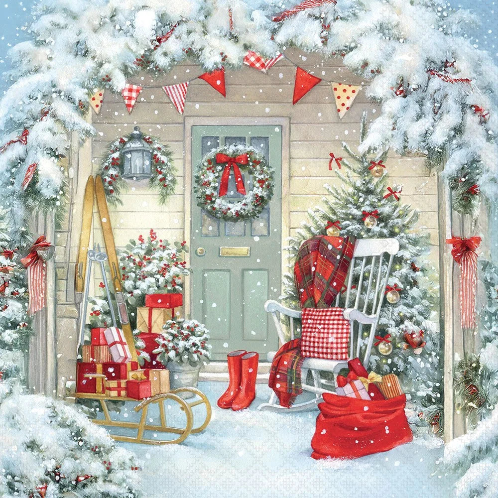 A festive Christmas porch scene with snow-covered trees and a house entrance. A green front door has a wreath and red bow, with red-and-white bunting, wrapped gifts, a sled, a rocking chair with a red plaid blanket, boots, and a sack of presents in the snow.
